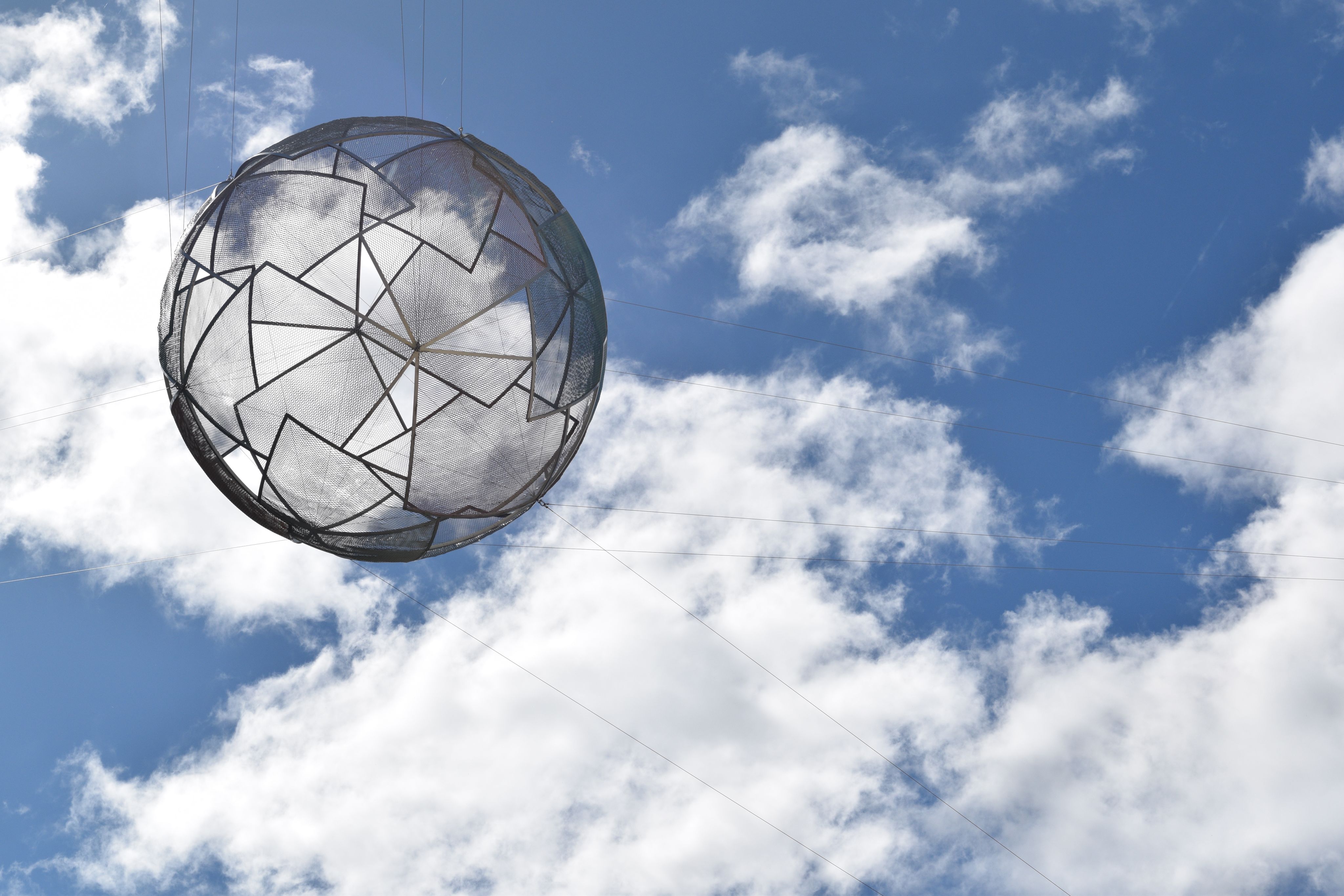 Geometric sphere suspended against blue sky with white, fluffy clouds.  