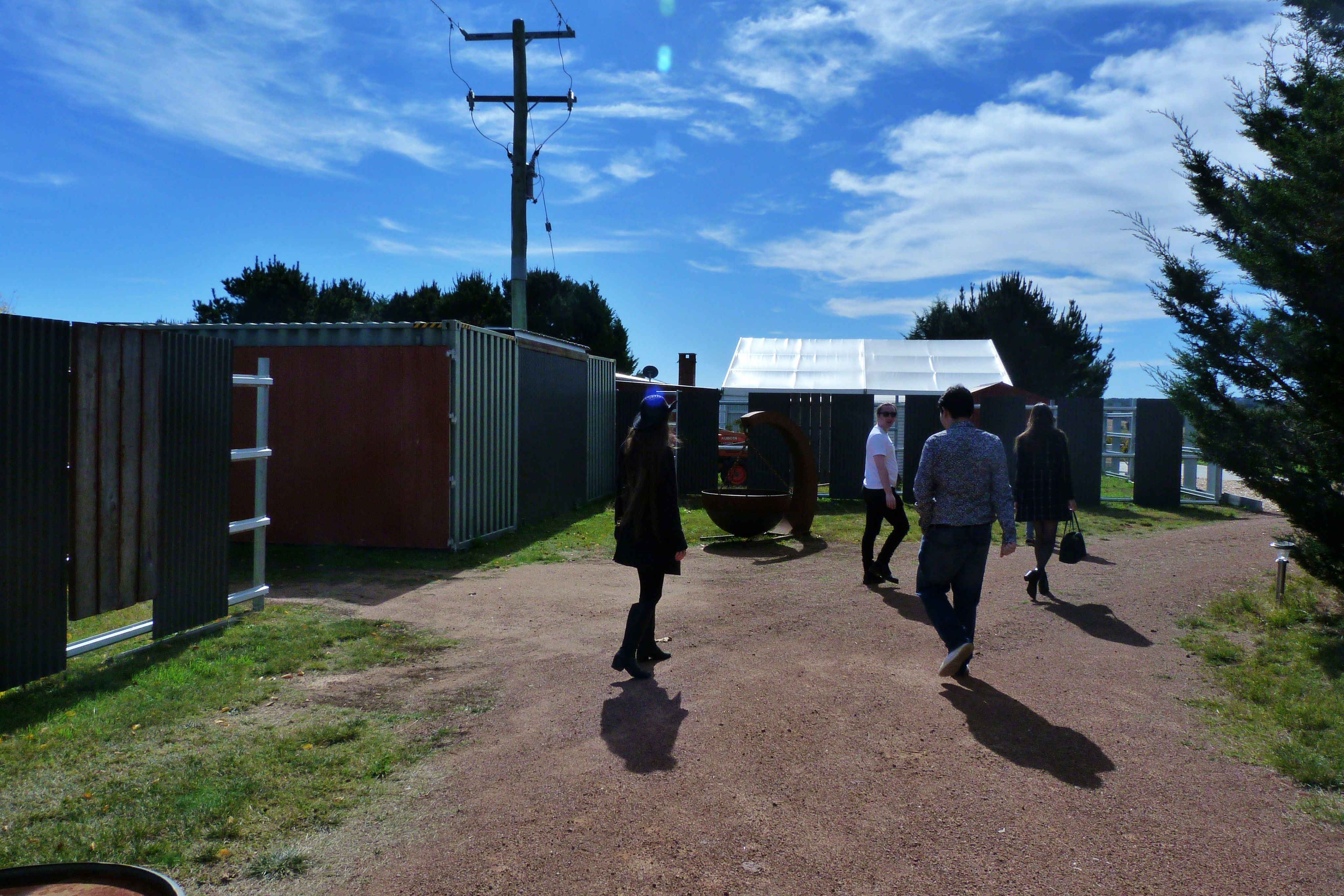 A group of people walking on a dirt road. There are framed by two metal sheds and a blue, cloud filled sky.