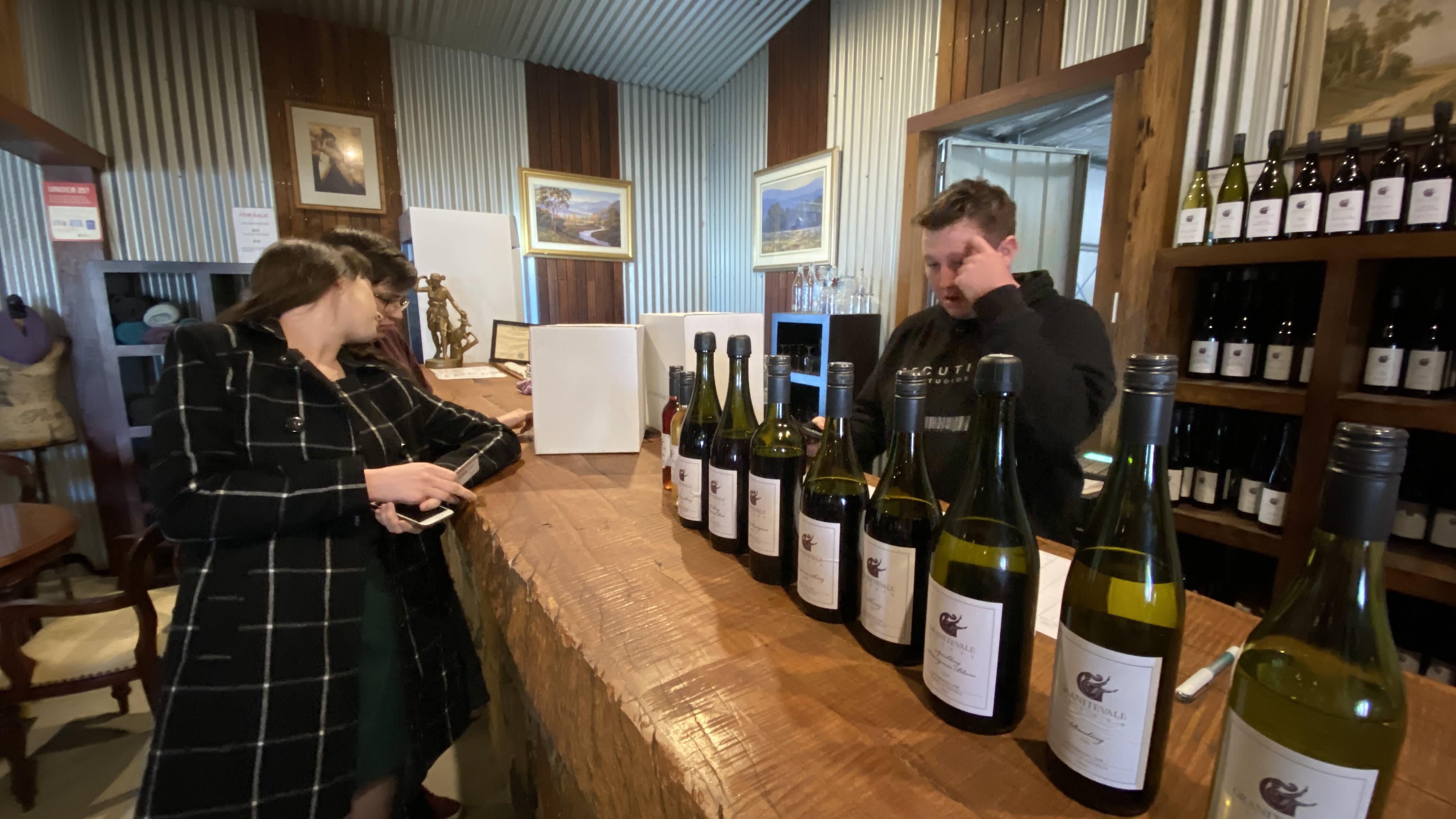 A pair of people stand on one side of a bar, with a man standing on the other. There are two white boxes between them, and a row of different wine bottles that line the table