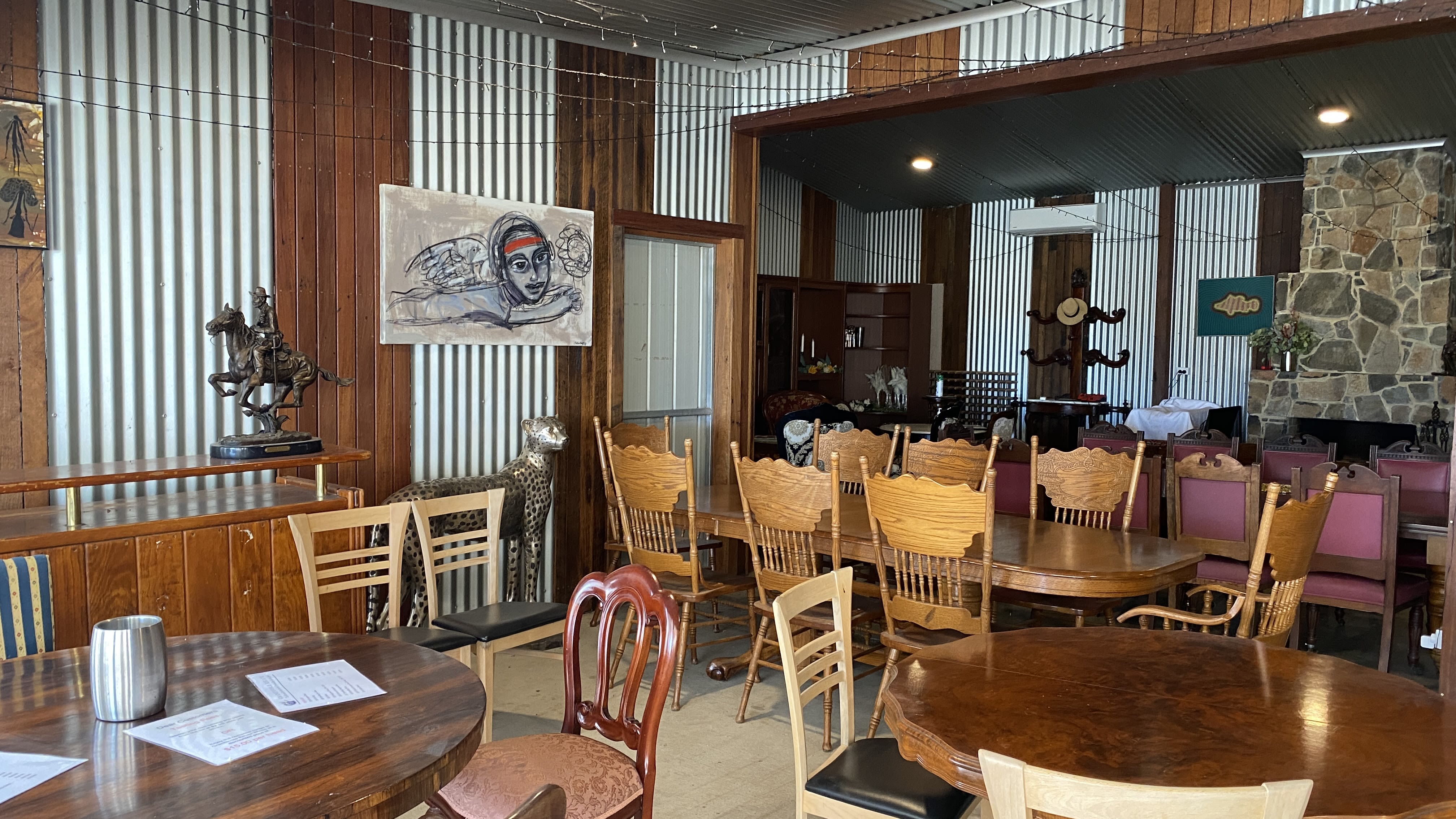 A collection of different kinds of tables and chairs inside a room with metal and wooden walls. Artworks hang on the walls, and in the corner is a stone fireplace.