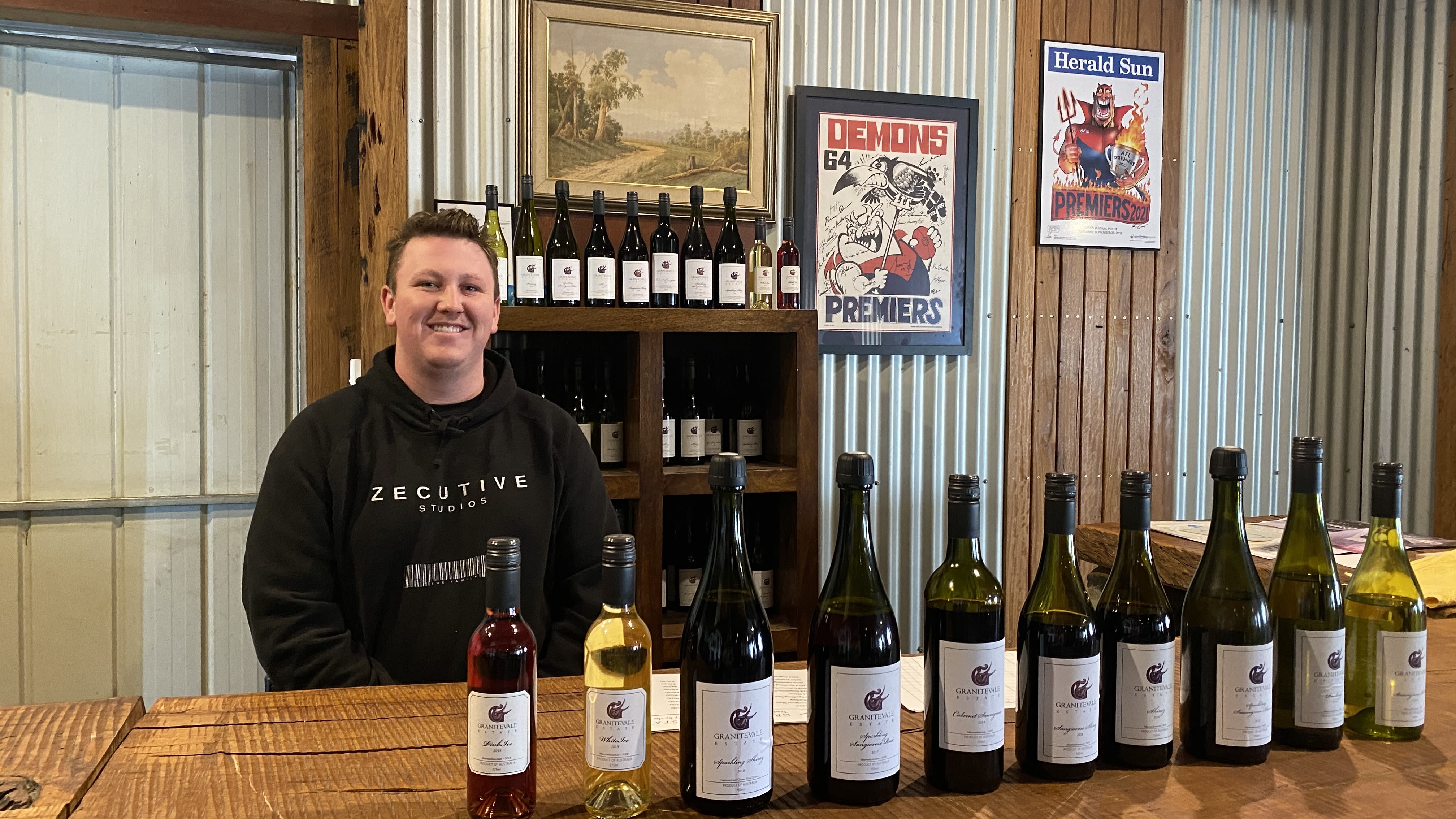 A man stands behind a bar, smiling at the camera. He wears a black hoodie. A collection of wines line the bar table.