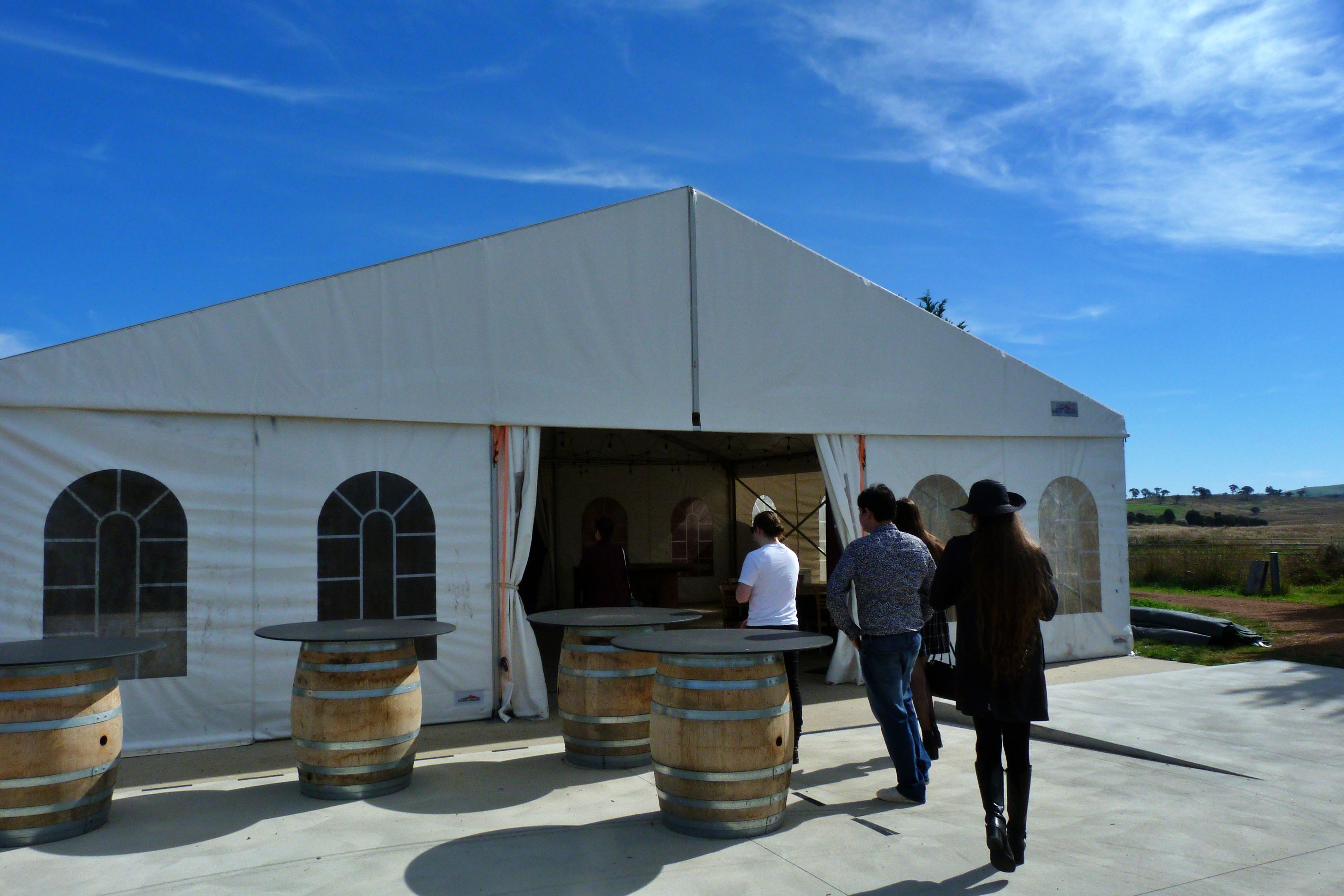 A group of people walk into a white tarp covered building. There are barrels standing vertical with slabs of metal on top, creating tables. In the distance are green vineyards and a blue sky.