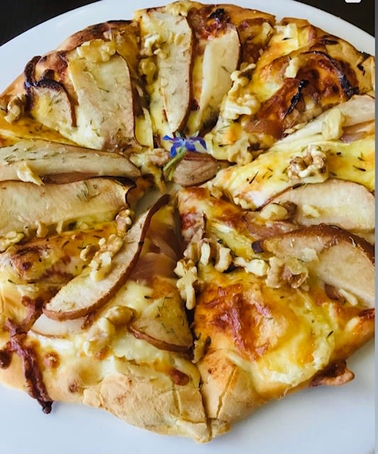 A close up of a pizza, it is topped with cheese, walnut pieces, and pear slices. A purple flower sits in the centre.