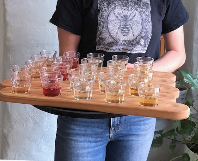 A person hold several wooden trays with small glasses of different coloured mead
