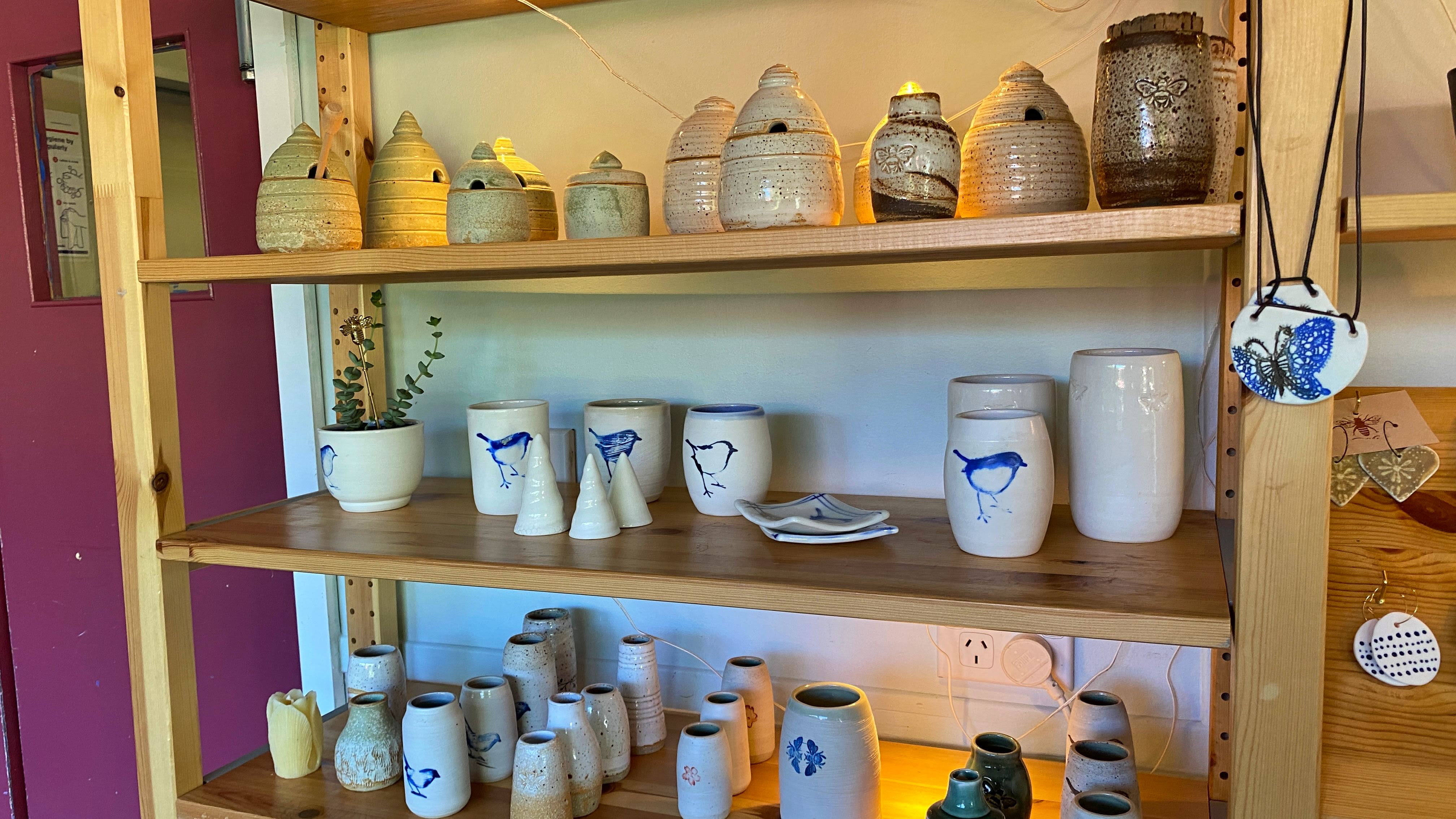 A row of shelves with different items of pottery on them. Many are beehive themed, or have a blue finch painted upon them. They are mostly mugs or honey storage containers.