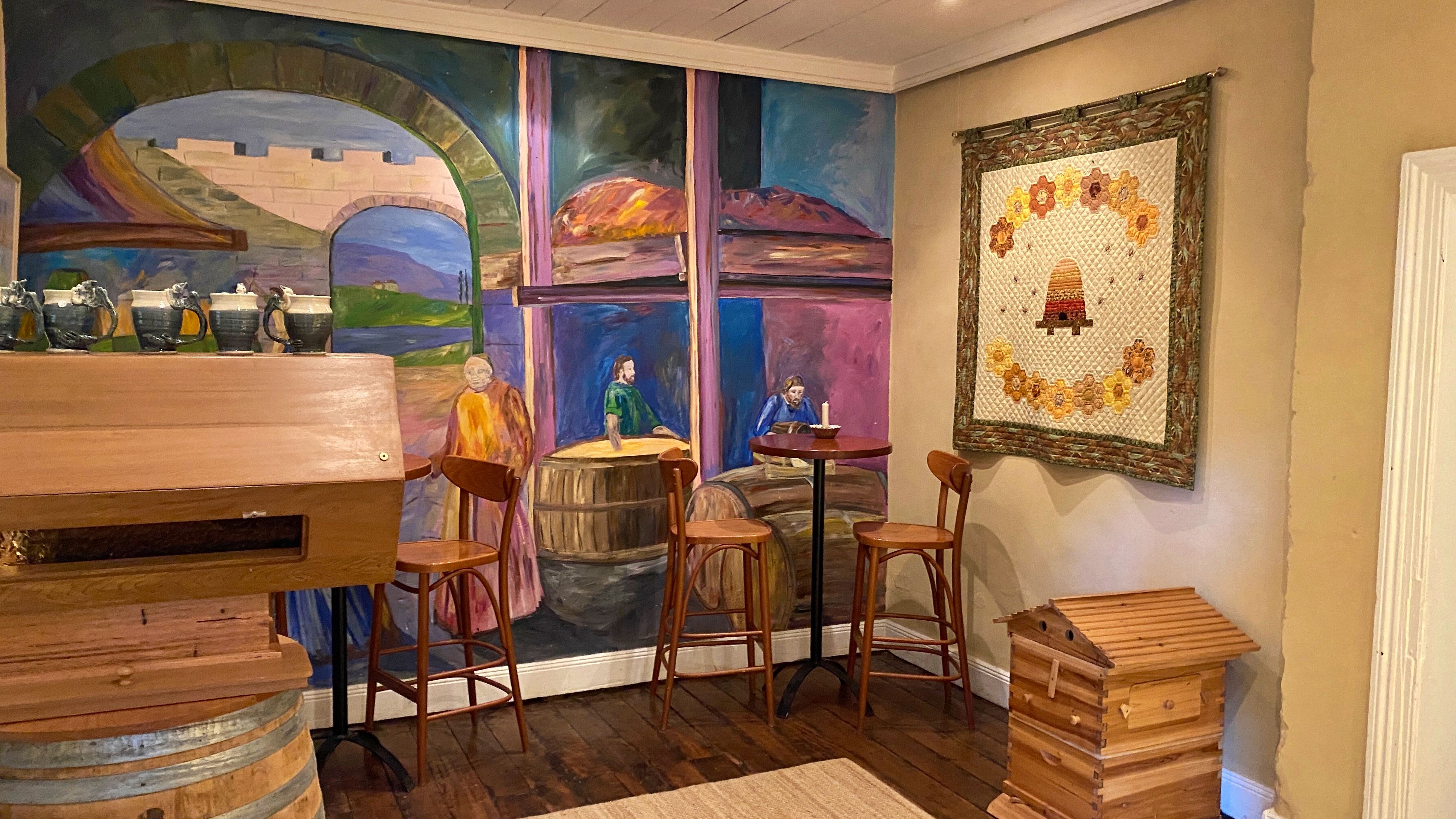 A wall mural showing people working in a barn is set behind a collection of tall tables and chairs, and a beehive.