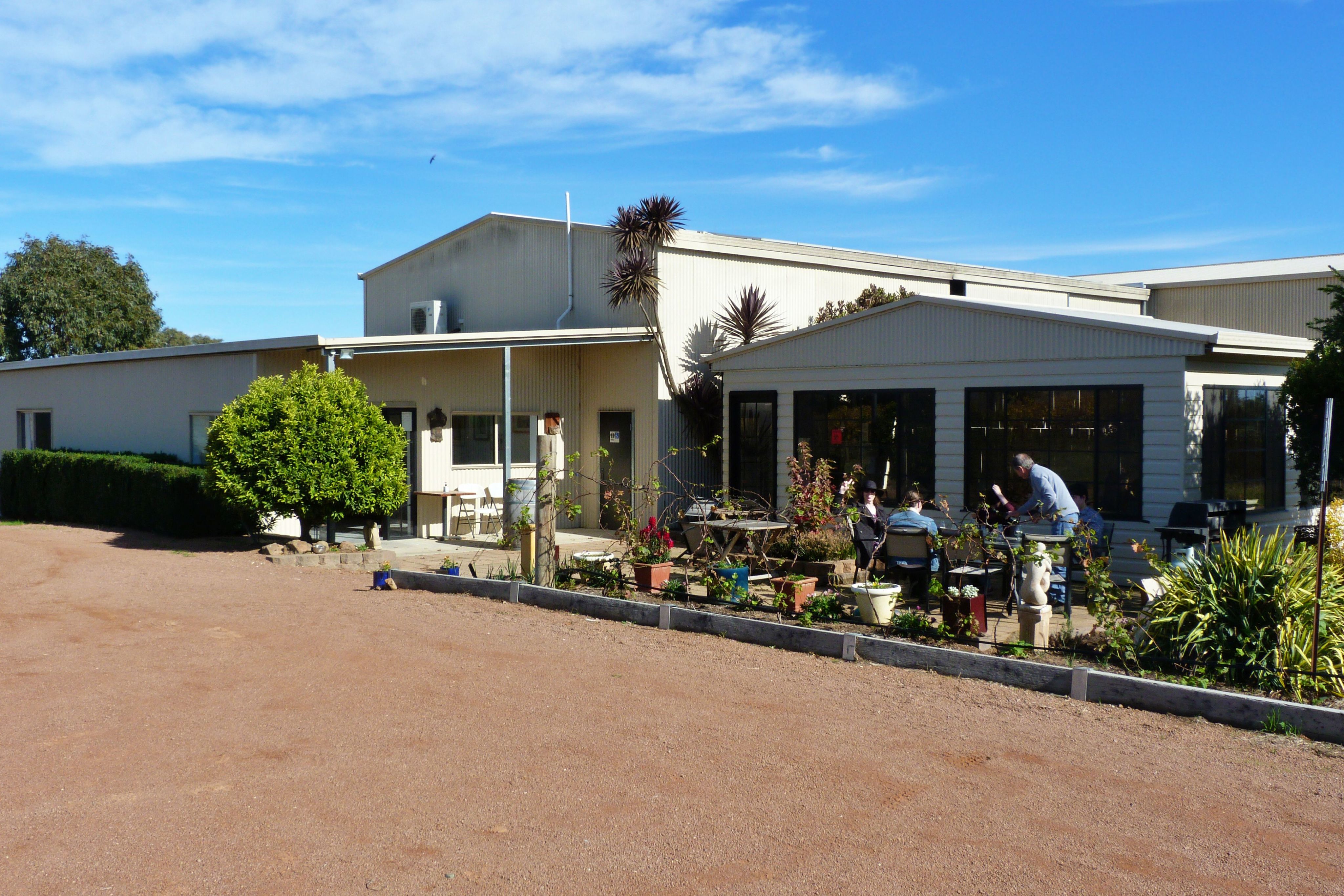 A white building surrounded by brown gravel. The building is lined by a garden with some shrubs and ornaments. People are gathering around a table.