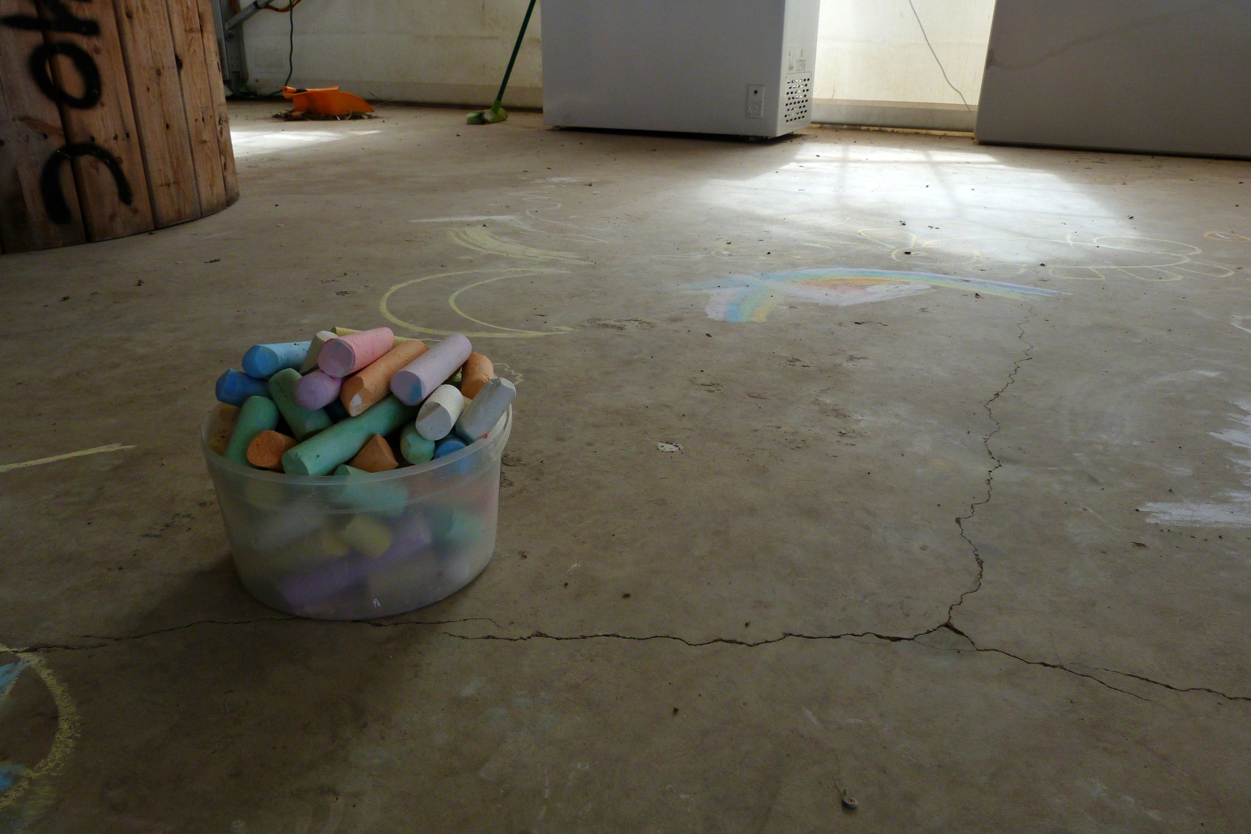 A bucket of multicoloured chalk sits surrounded by chalk artwork. It is inside a building, and is framed by tarp walls and a barrel.
