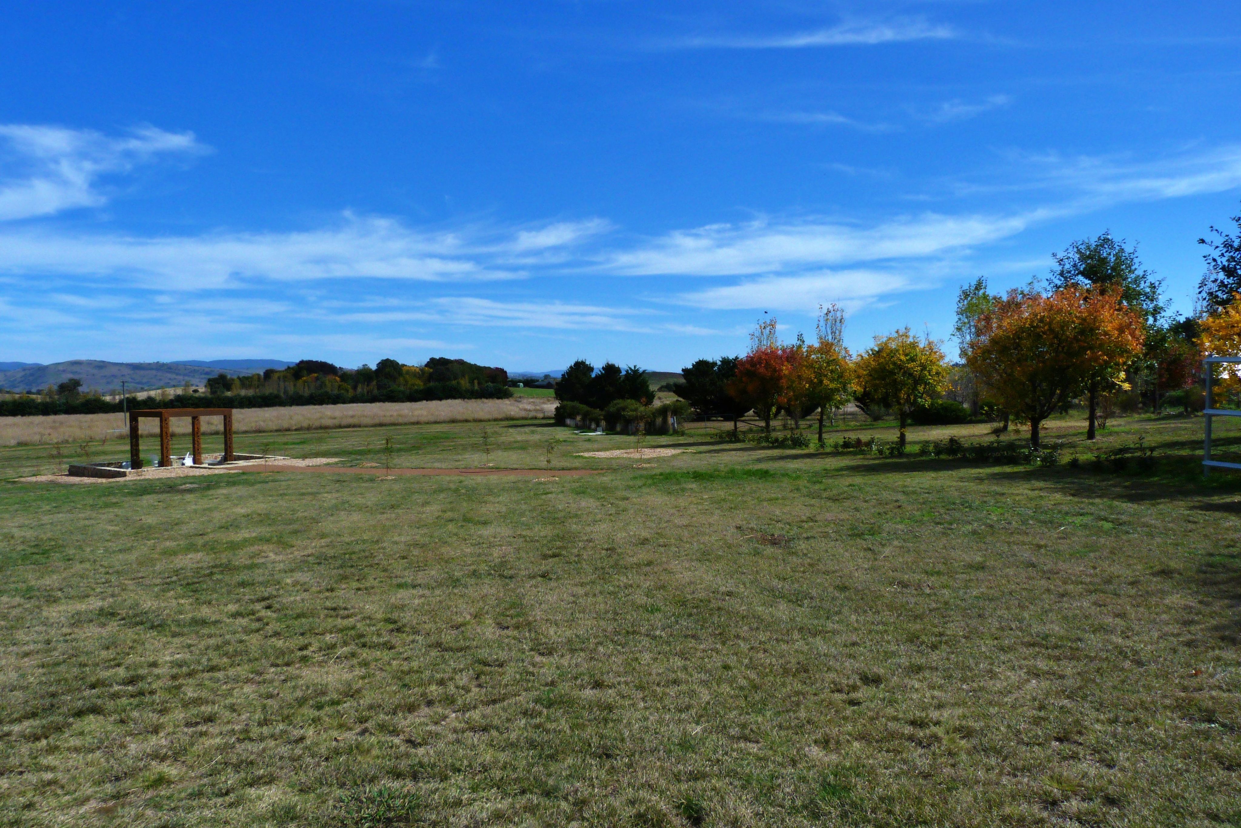 A field lined by trees turning orange and red due to Autumn. The grass is green, and sitting on the left is a metal gazebo style archway.
