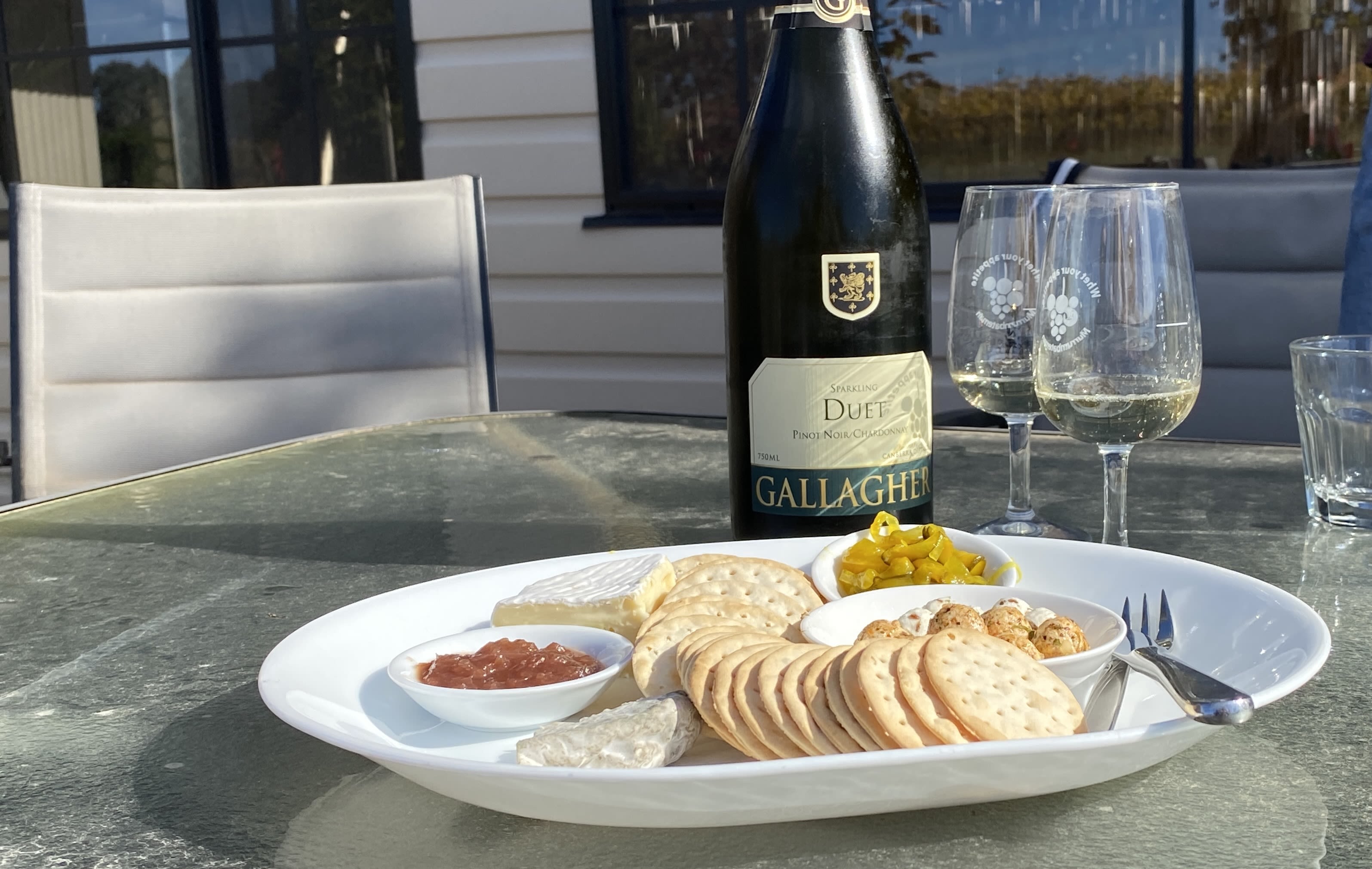 A cheese platter on a glass table. In the background is are two wine glasses filled a quarter length with white wine. A wine bottle labelled "Duet" stands by the glasses side.
