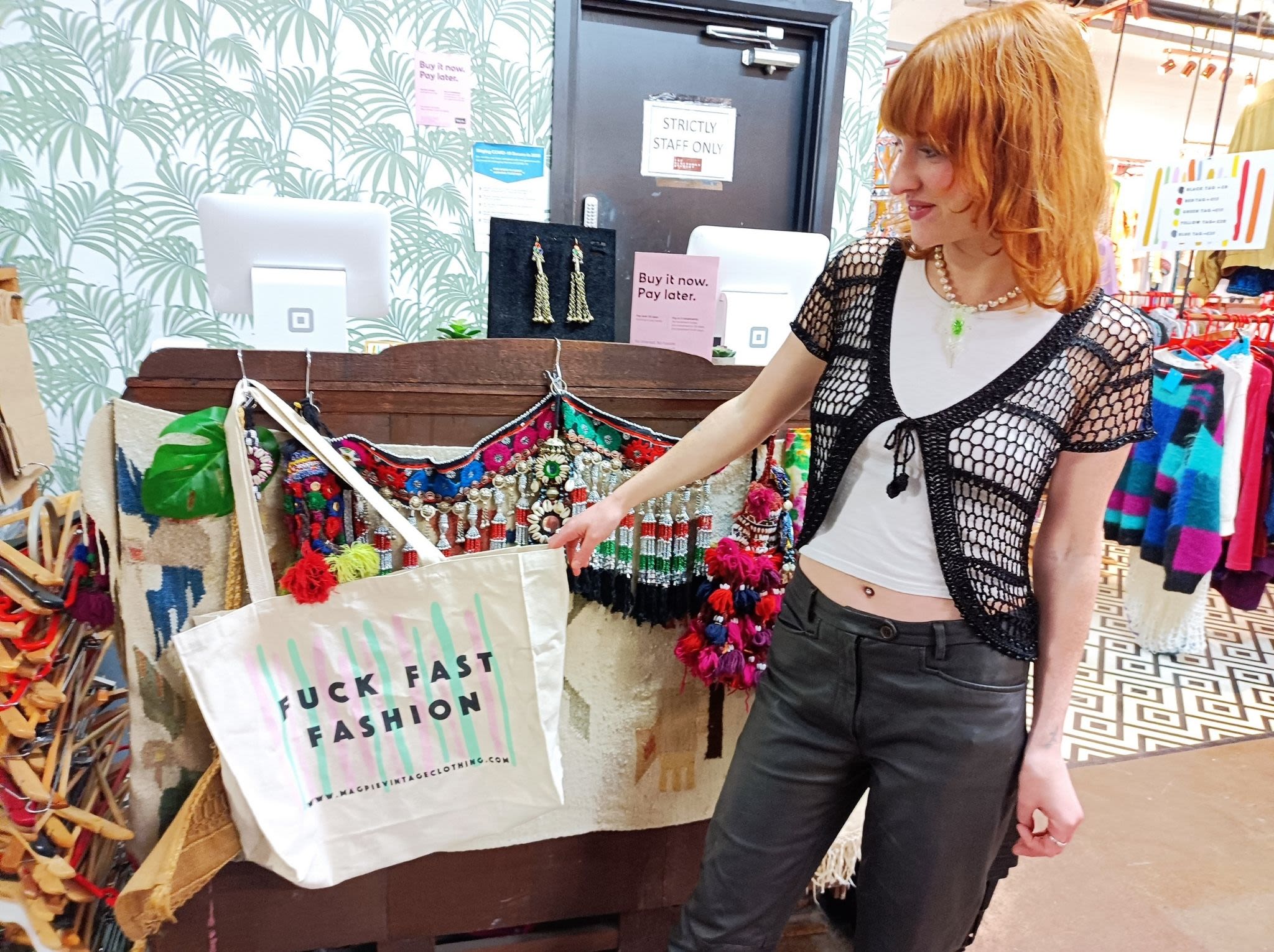A store employee wearing a black crotchet top over a white singlet, holds a bag out with the phrase "fuck fast fashion" printed on it in capital letters.
