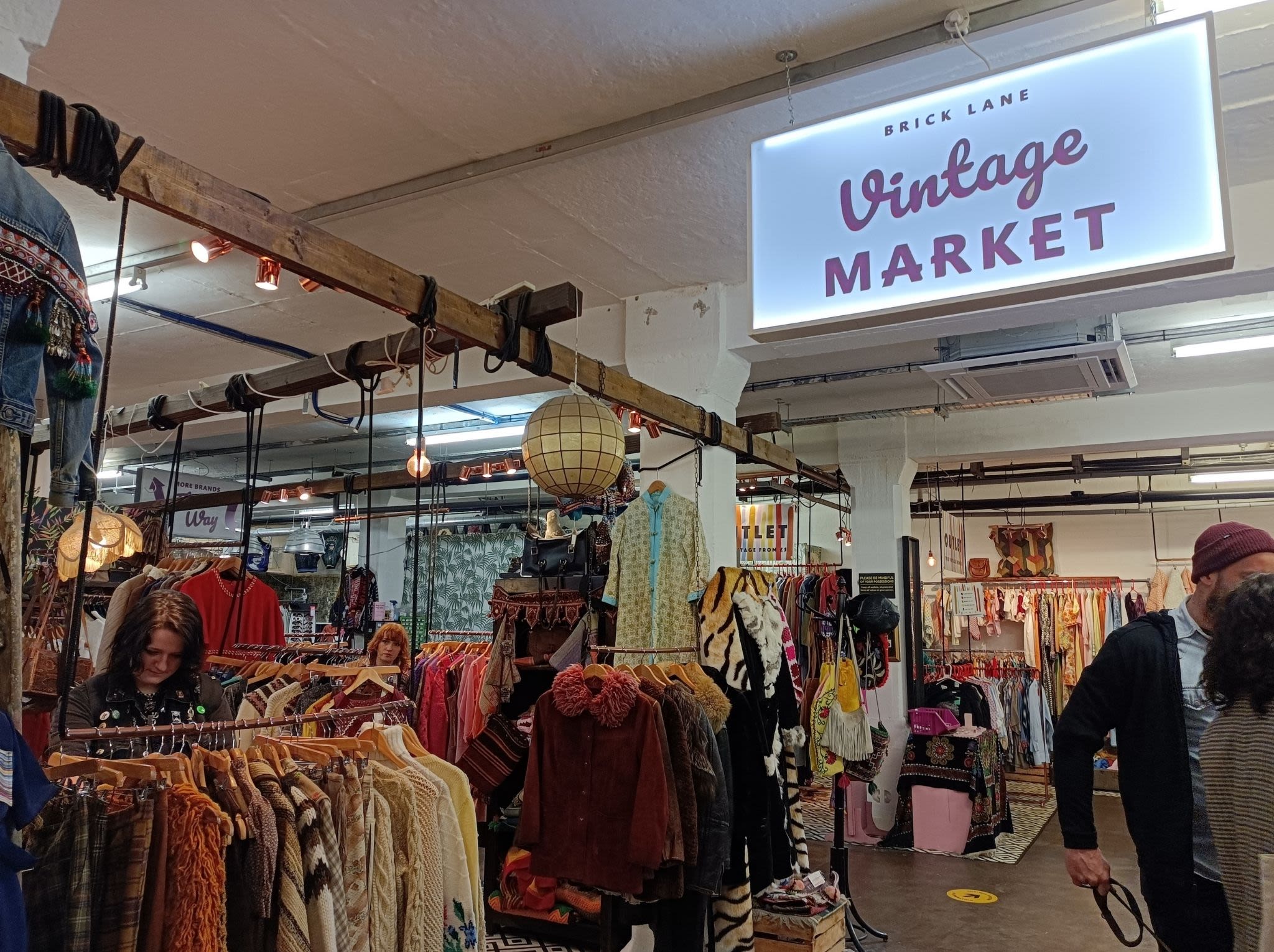 A wide shot of a few shoppers browse beneath a sign that says Vintage Market in purple above a wide shot of the market that is filled with colourful vintage jackets, dresses and accessories. 