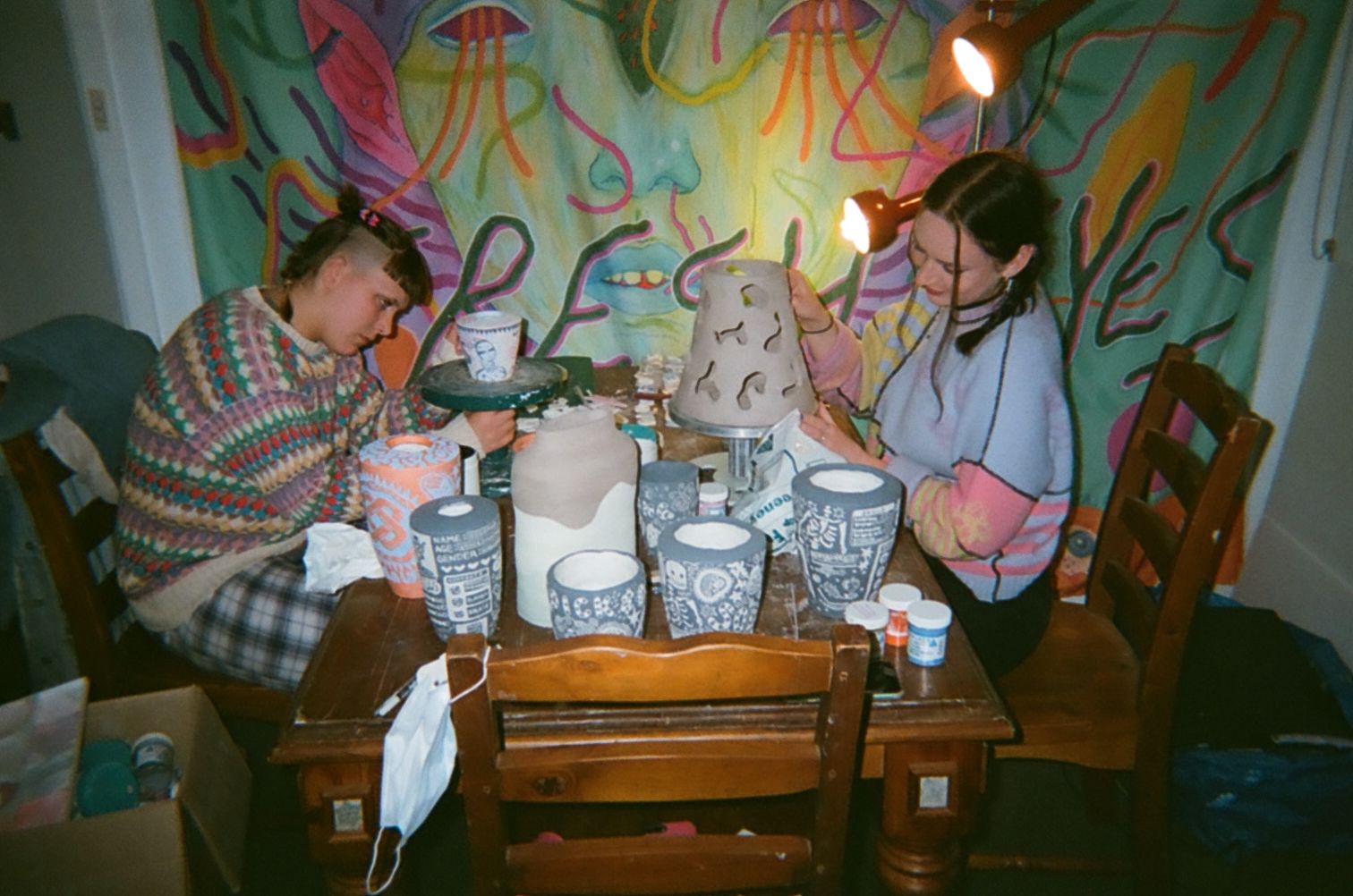 Emma and Eppie sit on opposites sides of their dining room able. The table is filled with ceramics, they are both working on individual pieces. Both in colourful jumpers behind a colourful backdrop