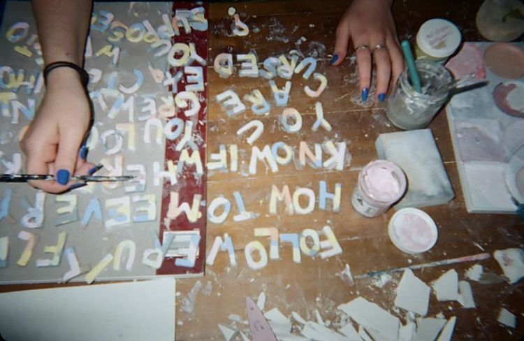 Emily works on colourful pottery letters on a bench. She has blue fingernails and is holding a paint brush
