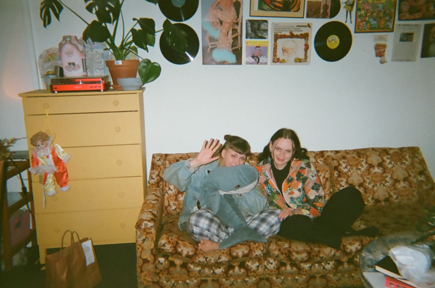 Eppie and Emily's living room. Their bright yellow couch can be seen, next to it a creamy yellow cabinet. A green plant sits on top of it, a doll hangs from the draws.