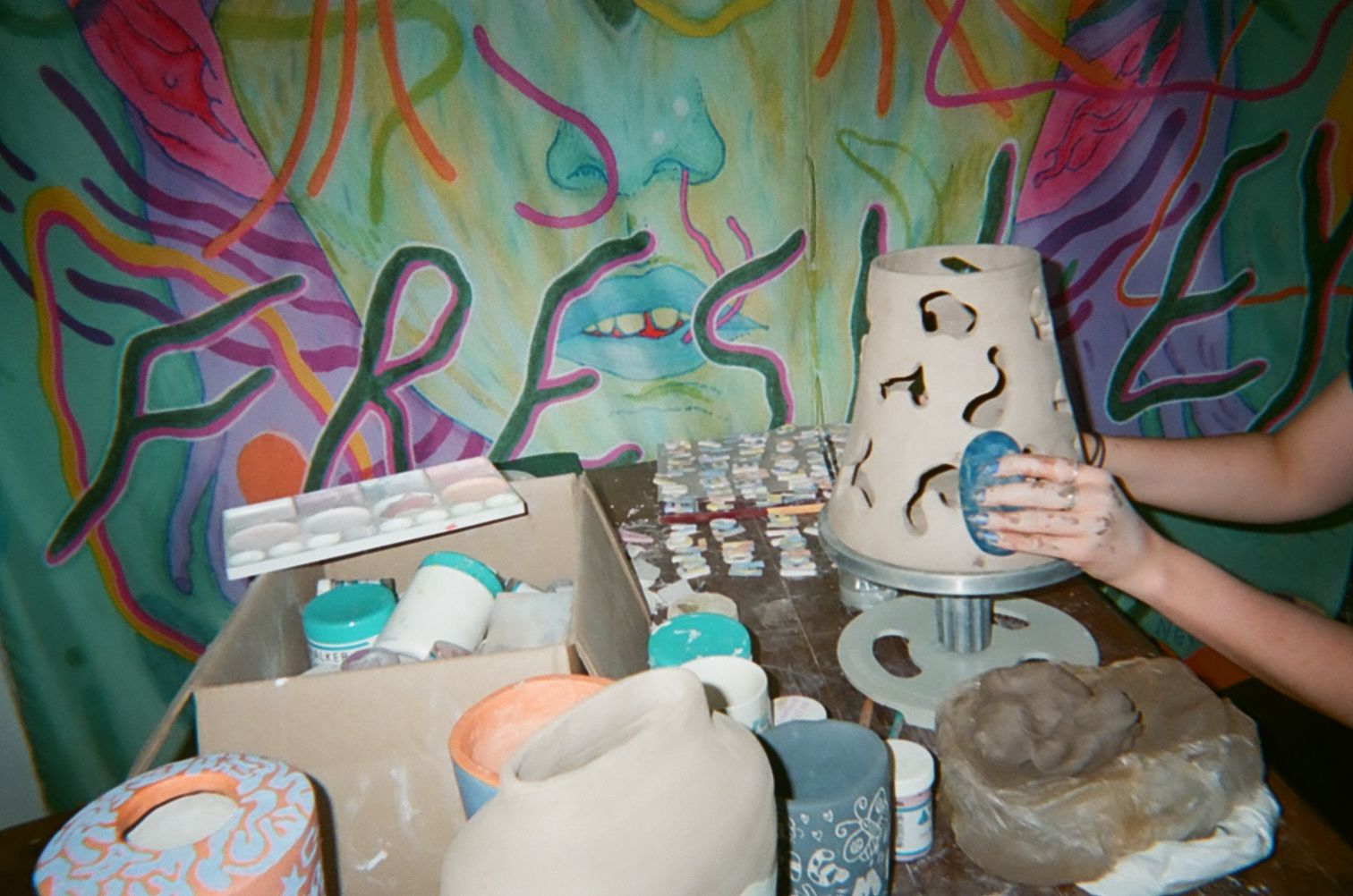 On a table covered in pottery, the hands of one of the pottery girls can be seen making touch up's to a ceramic lamp. The backdrop behind the desk is a colourful print