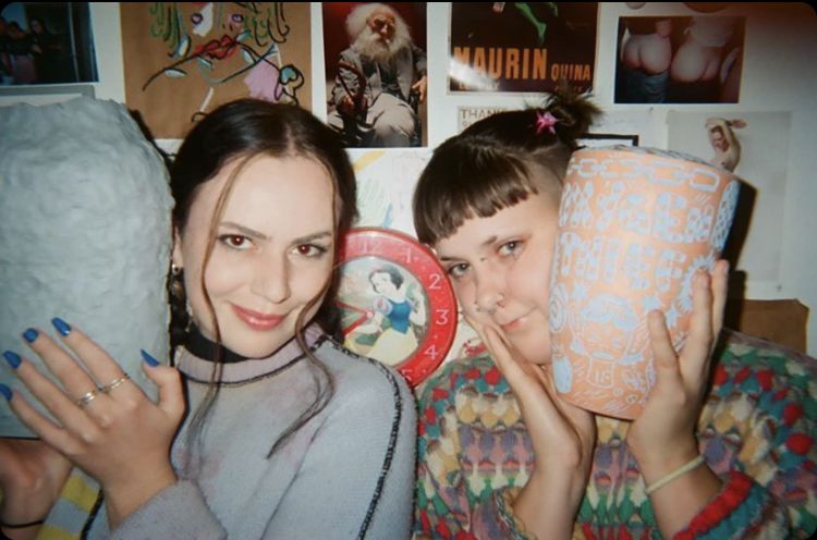 Emma in blue jumper holds blue vase, Eppie in colourful jumper holds orange and blue vase.